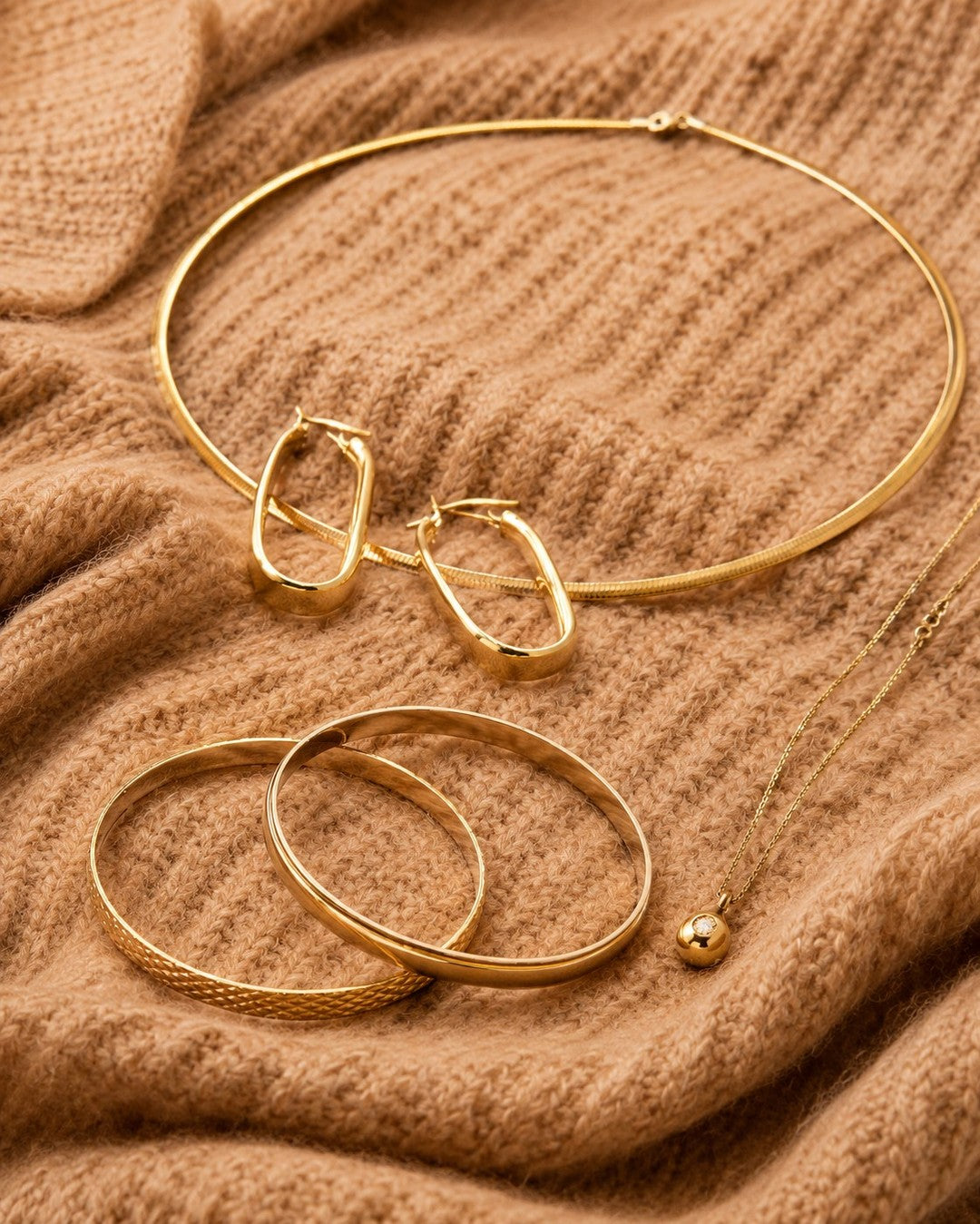 Gold-tone necklaces, hoop earrings and bangles arranged on warm textured fabric.